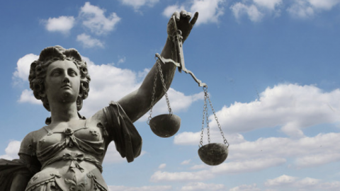 Die Statue der Justitia steht erhaben und hält die Waage in einer Hand. Der Himmel ist blau mit Wolken.