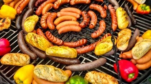 Auf einem Grill sind verschiedene Wurstsorten und Brötchen angeordnet, während Menschen im Hintergrund entspannt stehen.