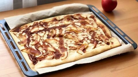 Apfelpfannkuchen auf Blech, gebacken mit Zimt und Apfelscheiben, steht auf einem Tisch neben einem roten Apfel.