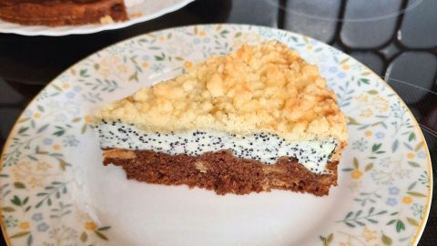 Ein Stück Quark-Mohn-Kuchen liegt auf einem blumendekorierten Teller, umgeben von einem weiteren Kuchen auf einer weißen Servierplatte.