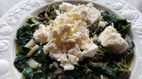 Ein frischer griechischer Wildkräutersalat aus dunklem Blattgemüse liegt auf einem kunstvollen Teller, garniert mit zerbröckeltem Feta.