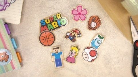 Bunte Sticker, darunter Super Mario, Basketball und eine Blume, liegen auf einem hellen Untergrund neben Bürobedarf.