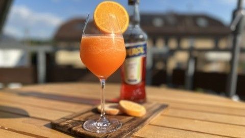 Ein kristallklarer Aperol Spritz in einem hohen Glas mit einer Orangenscheibe steht auf einem Holzbrett im Freien.