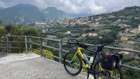 Ein Fahrrad steht auf einem Aussichtspunkt, mit Blick auf grüne Hügel und italienische Landschaft, während Wolken vorbeiziehen.