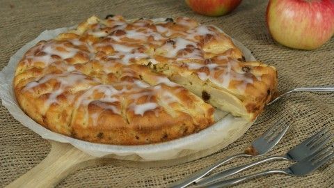 Ein gedeckter Apfelkuchen mit Rum-Rosinen, leicht glasiert, steht auf einem Holzbrett, umgeben von frischen Äpfeln.