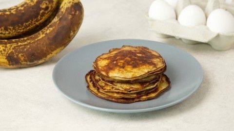 Auf einem grauen Teller stapeln sich goldbraune Pfannkuchen, während überreife Bananen neben frischen Eiern liegen.