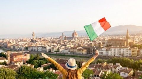 Eine Frau mit einem Hut hält eine italienische Flagge hoch und blickt auf die Stadt Florenz bei Sonnenuntergang.