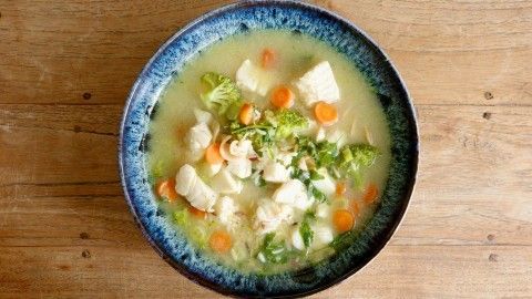 Eine bunte Fischsuppe mit Brokkoli, Karotten und zartem Fisch in einer blauen Schüssel, auf einer Holzoberfläche serviert.