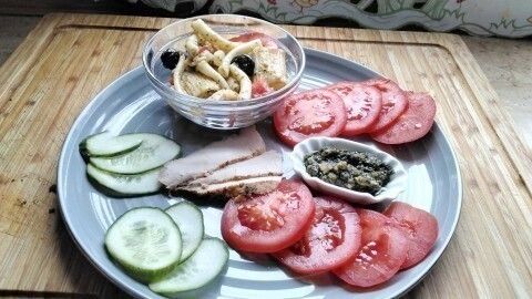 Ein bunter Nudelsalat mit Pesto und Oliven liegt in einer Schüssel, umgeben von frischen Tomaten und Gurkenscheiben.