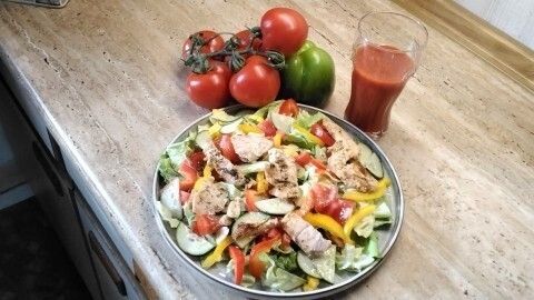 Frischer, bunter Salat mit Lachs liegt auf einem Tisch, umgeben von Tomaten und Paprika sowie einem Glas Saft.