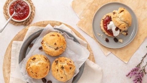 Auf einem Teller liegen goldbraune Scones mit Rosinen, die mit Clotted Cream und Marmelade serviert werden.