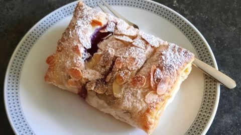 Delikate Blätterteigtasche mit Heidelbeeren liegt auf einem Teller, verziert mit Puderzucker und Mandelsplittern.