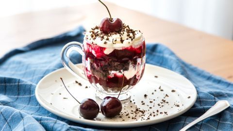 Ein Glas Schwarzwälder-Kirsch-Dessert steht auf einem Teller, umgeben von Kirschen und einer blauen Tischdecke.