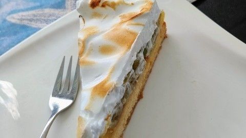 Ein Stück Stachelbeerkuchen mit flambierter Schaumdecke liegt auf einem weißen Teller, umgeben von einem blauen Tisch mit maritimem Design.