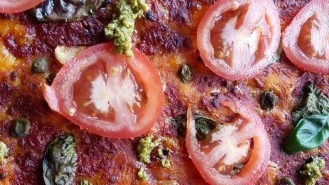 Eine vegane Pizza liegt appetitlich auf einem Teller, belegt mit frischen Tomatenscheiben, Basilikum und grünem Pesto.