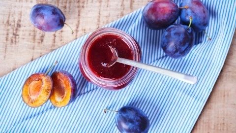 Ein Glas Pflaumenmarmelade steht auf einem blauen Tuch, umgeben von frischen Pflaumen, auf einer Holzoberfläche.