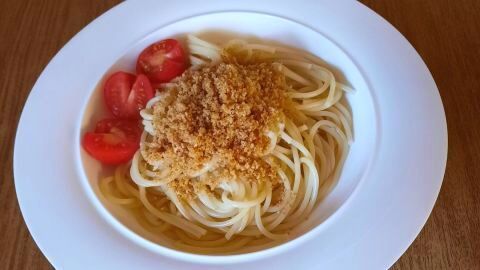 Spaghetti mit goldbraunen Semmelbröseln und geschnittenen Tomaten auf weißem Teller, präsentiert auf holzfarbener Oberfläche.