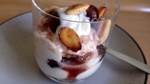 Ein Glas Kirsch-Dessert mit Joghurt ist dekoriert mit Sahne und gerösteten Mandeln, platziert auf einem grauen Teller.