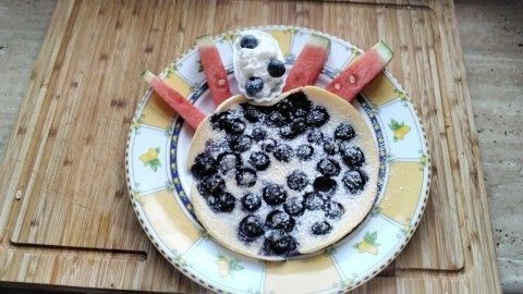 Ein großer Heidelbeerpfannkuchen liegt auf einem bunten Teller, dekoriert mit Sahne und Wassermelonenstücken.