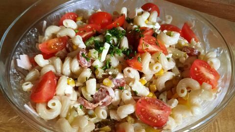 Ein Nudelsalat mit Tomaten, Mais und Kräutern ruht in einer Glasschüssel auf einem Holztisch; ein Holzlöffel liegt daneben.