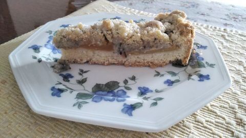 Ein Pfirsichkuchen mit Nussstreuseln liegt auf einem blauen Blumen-Servierteller, sanft angestrahlt von Tageslicht.