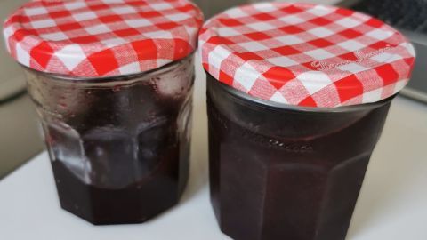 Zwei Marmeladengläser mit rot-weiß karierten Deckeln stehen auf einem weißen Tisch in einer modernen Küche.