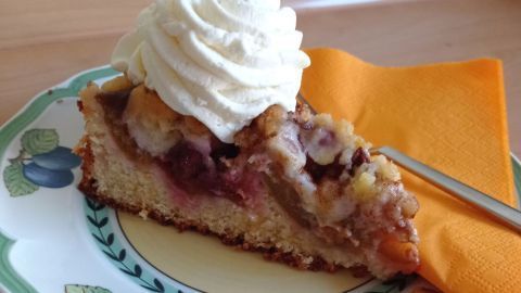 Ein Stück Pflaumenkuchen mit Sahnehaube auf einem dekorativen Teller, daneben eine orange Serviette auf einem Holztisch.