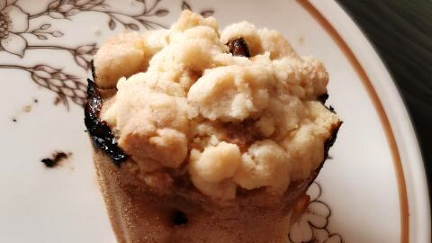 Muffin mit Streuseln steht auf floralem Porzellanteller, umgeben von Holzoberfläche. Keine sichtbare Schrift vorhanden.