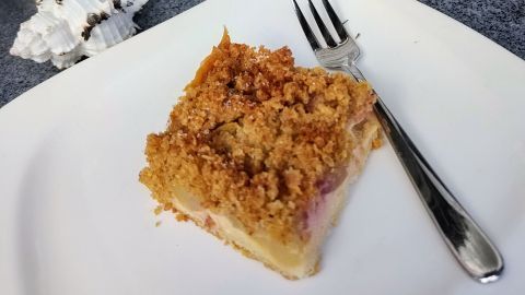 Ein Stück Obstkuchen mit Haferflockenstreuseln liegt auf einem Teller neben einer Gabel, dekoriert mit Pflanzen und Kürbissen.