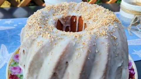 Ein Gugelhupf mit Zuckerguss steht auf einem blumigen Teller, umgeben von Pflanzen auf einem Tisch im Garten.