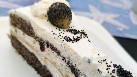 Ein Stück Mohn-Marzipan-Torte auf einem Teller zeigt Schichten von Sahne und Mohn, im Hintergrund ein dekoriertes Glas.