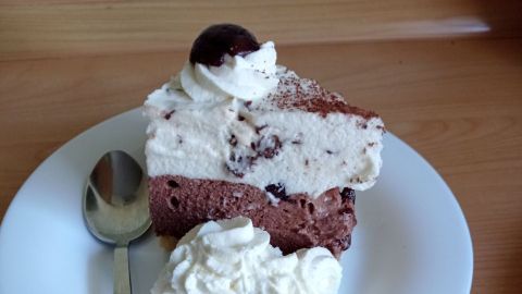 Ein Stück Paradiescreme-Schnitte mit Kirsche obendrauf, serviert auf einem Teller mit Sahne und Löffel, auf Holztisch.