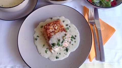 Kabeljau auf Teller in Zitronen-Kapern-Soße, mit Petersilie bestreut, daneben Kartoffeln, Salat und separate Soßenschale.