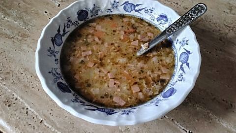 Eine keramikblaue Schüssel mit bunter Suppe, Kartoffelstücken und Wurstwürfeln steht auf einem Marmortisch, ein Löffel darin.