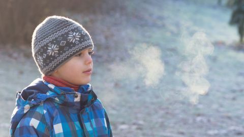 Ein Kind mit Mütze atmet im Freien; der Atem ist sichtbar in der kalten Winterluft auf einem frostigen Rasen.