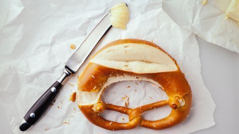 Butterbrezel auf Papier, daneben ein Messer mit Butter, umgeben von kleinen Krümeln.