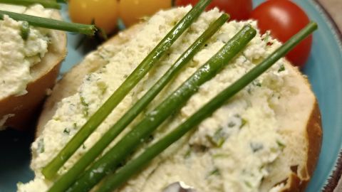 Brötchenhälfte mit Kräuterfrischkäse bestrichen, belegt mit Schnittlauch; im Hintergrund Tomaten auf blauem Teller. Datum: 26. Sept. 2024, 19:15.