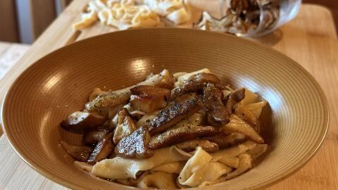 Tagliatelle mit Steinpilzen in einer braunen Schüssel auf einem Holztisch; hinten liegen rohe Nudeln und ein Glas mit Steinpilzen.