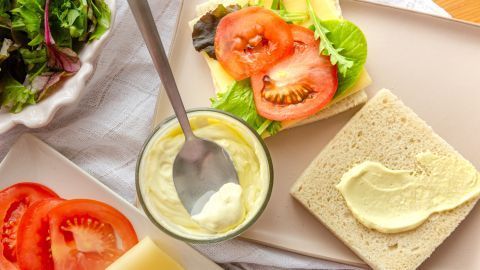 Ein Glas Mayonnaise ohne Ei mit Löffel steht neben belegtem Brot mit Tomaten und Salat auf Teller, umgeben von frischem Gemüse.