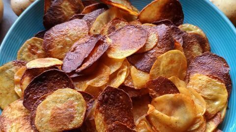 Kartoffelchips in einer blauen Schale, knusprig gebacken, umgeben von rohen Kartoffeln. Text: 6. Okt. 2024, 14:48.