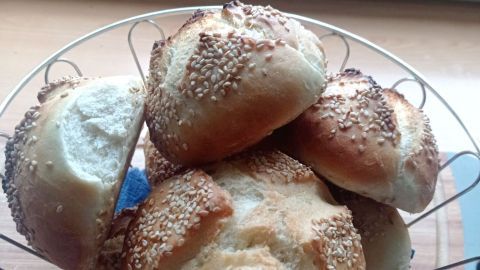 Sesambrötchen liegen frisch gebacken in einem Metallkorb auf einer Holzplatte vor einem Fenster mit Blick ins Grüne.