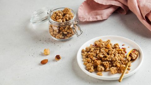 Ein Glas und ein Teller mit knusprigem Müsli stehen auf einem hellen Tisch; daneben liegen eine Nuss und ein rosafarbenes Tuch.