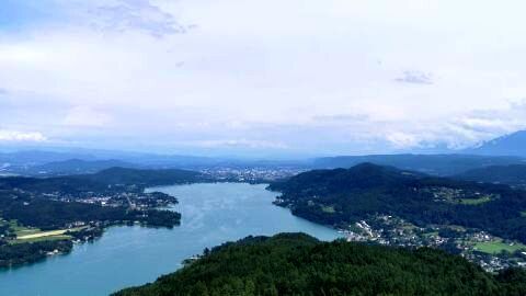 Ein weiter See erstreckt sich durch bewaldete Hügel, während am Horizont eine Stadt unter einem blauen Himmel liegt.