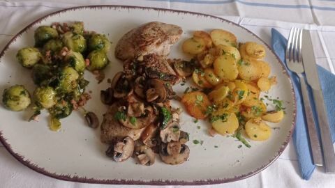 Auf einem Teller liegen Waidmanns-Filets mit Rosenkohl und Bratkartoffeln, getoppt mit frischen Kräutern und Pilzen auf den Filetscheiben.