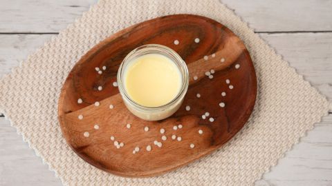 Ein Glas mit handgemachter Creme steht auf einem Holztablett, umgeben von Bienenwachs, auf einer strukturierten Tischdecke.