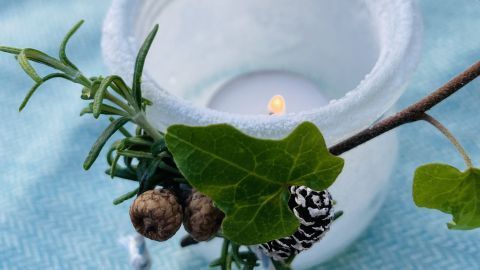 Ein transparentes Windlicht mit einem flackernden Licht steht auf einem hellblauen Tuch, umgeben von grünen Zweigen und Beeren.