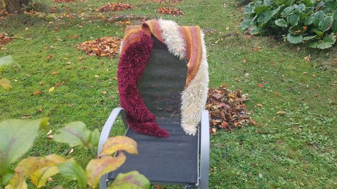 Eine gestrickte, herbstliche Stola liegt dekorativ auf einem Gartenstuhl, umgeben von buntem Herbstlaub auf grüner Wiese.