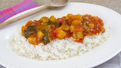 Teller mit Ratatouille auf Langkornreis, garniert mit einem Holzlöffel, auf einem Holztisch mit farbiger Serviette.