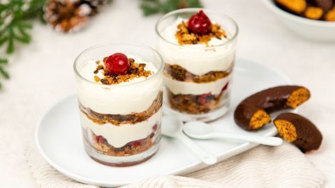 Zwei Gläser Lebkuchen-Tiramisu mit Schichten aus Creme und Lebkuchen, dekoriert mit Kirschen, auf einem Teller mit Löffeln.