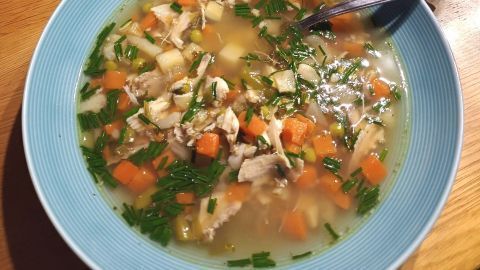 Gemüsesuppe in einer blauen Schale mit Huhn und Gemüsewürfeln; daneben ein Topf und Brötchen auf einem Holzbrett.